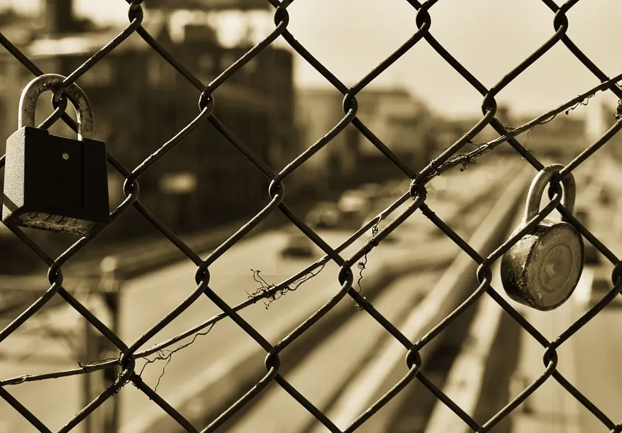 Two padlocks clipped onto a chain-link fence, in sepia, with a blurred street behind them