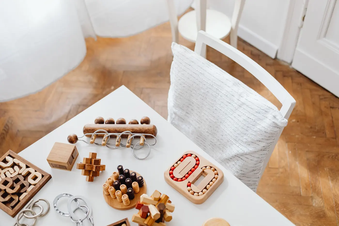 Wooden brain-teaser puzzles, metal ring puzzles and peg games spread across a white table beside a chair