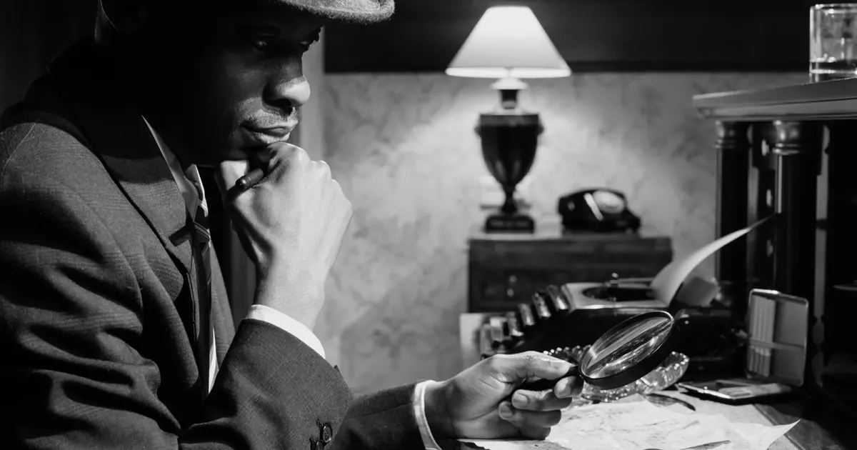 Black-and-white photo of a man in a fedora studying papers through a magnifying glass at a desk with a typewriter and lamp
